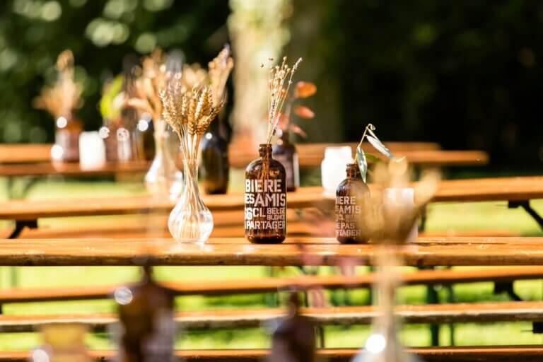 Cette image met en valeur une décoration de table élégante et champêtre pour un mariage dans les Vosges. Des bouteilles de bière ambrées, réutilisées en vases, accueillent des épis de blé et des fleurs séchées, ajoutant une touche naturelle et authentique à la scénographie. L'ensemble s'accorde parfaitement avec les bancs en bois sous un soleil doux, dans un cadre extérieur verdoyant. Cette photo capture à la perfection l'esprit bohème et chaleureux d'un mariage en plein air, où simplicité et élégance se rencontrent.