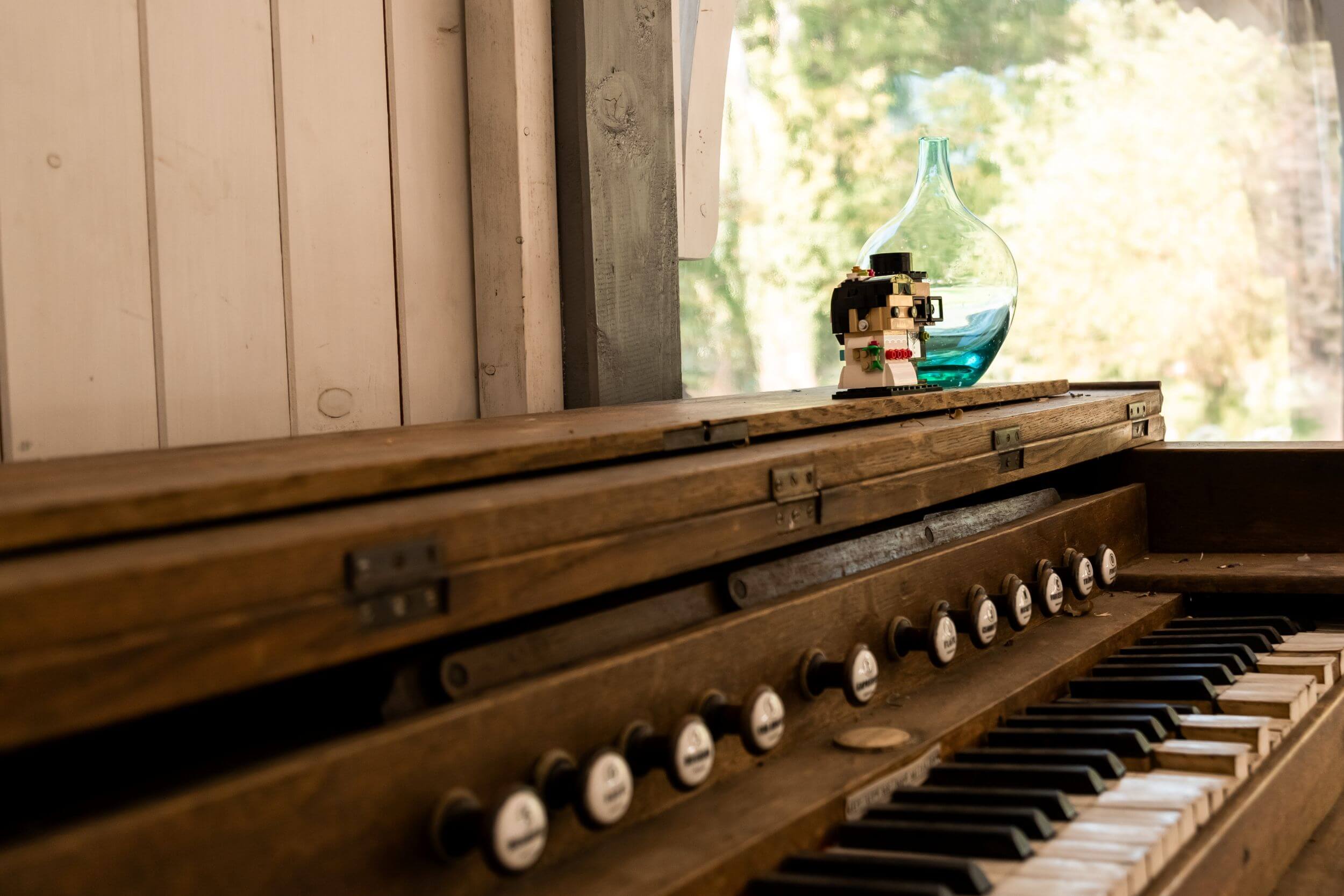 L'authenticité et l'originalité prennent vie sur cette photo où un orgue ancien, en bois patiné, est sublimé par des éléments décoratifs uniques. En premier plan, un objet LEGO stylisé apporte une note ludique et décalée, tandis qu'un vase en verre bleu illumine l'arrière-plan. Cette image symbolise l'harmonie entre tradition et modernité, idéale pour un mariage atypique où chaque détail raconte une histoire. Mariage vintage, photographe mariage Vosges, décoration orgue ancien, mariage personnalisé, détails uniques mariage.